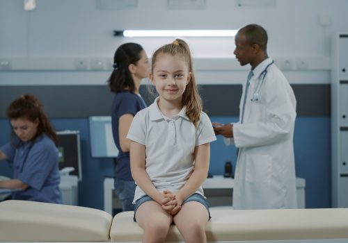 Portrait of little girl on bed waiting to begin checkup exam in medical cabinet with doctor. Small kid with illness looking at camera and getting ready for checkup visit appointment in office.