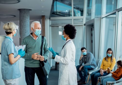 mature man and healthcare workers communicating in a hallway at medical clinic while wearing protective face masks.